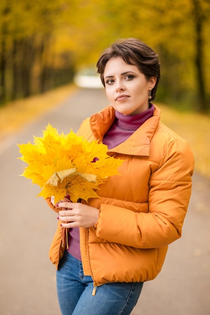 Piękna Brunetka Kobieta Pozuje W żółtej Kurtce W Jesiennym Parku Z Bukietem Jesiennych Liści I Patrzy W Kamerę