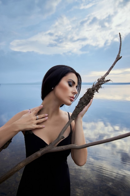 Piękna brunetka kobieta w czarnej sukni pozuje w morskim jeziorze na tle błękitnego nieba. Seksowna kobieta z długimi włosami i piękny makijaż na twarzy