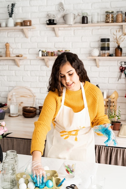 Piękna brunetka kobieta w żółtym swetrze i szarym fartuchu, kolorując pisanki na niebiesko w kuchni