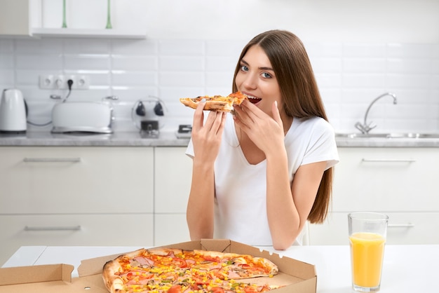 Piękna brunetka siedzi w kuchni i je pizzę