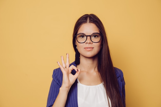 Piękna Brunetka W Okularach Z Długimi Włosami Ubrana W Granatowy Garnitur, Pokazuje Znak Ok, Wygląda Pewnie, Na żółtym Tle Z Miejsca Kopiowania. Koncepcja Ludzi Sukcesu.