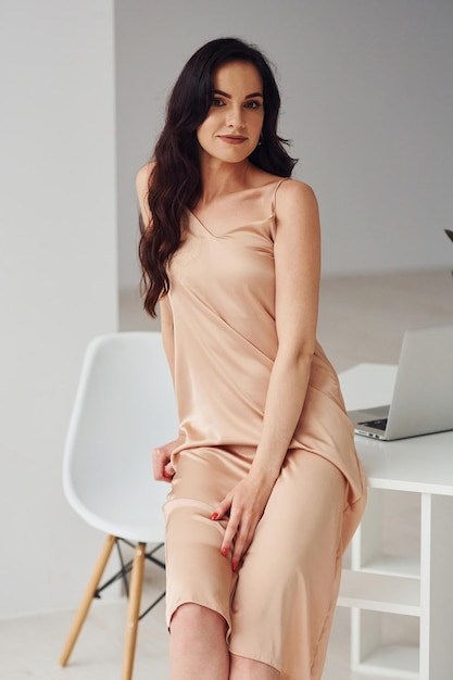 Piękna brunetka z kręconymi włosami w luksusowych ubraniach, stojąca przy stole z laptopem w domu w ciągu dnia