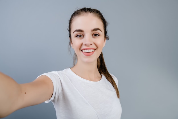 Piękna brunetki dziewczyna jest ubranym białą koszula robi selfie odizolowywającemu nad popielatym