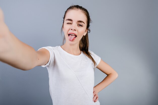 Piękna brunetki dziewczyna jest ubranym białą koszula robi selfie odizolowywającemu nad popielatym