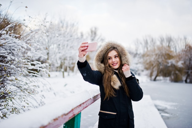 Piękna brunetki dziewczyna w zimy ciepłej odzieży. Modeluje na kurtki zimowej przeciw zamarzniętemu jezioru przy parkiem robi selfie na telefonie.