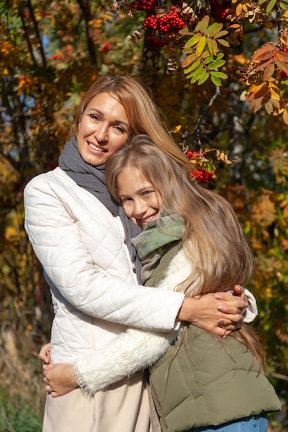 Piękna caucasian mama ściska córki w futerkowych hełmofonach na tle kolorowi jesieni drzewa. Koncepcja weekendu rodzinnego, miłość, relaks na świeżym powietrzu, zabawa, ludzie, pora roku, ciepła pogoda, malownicze lasy