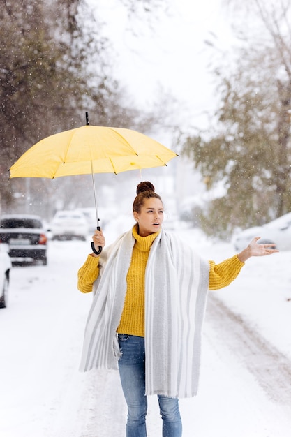 Piękna ciemnowłosa dziewczyna ubrana w żółty sweter, dżinsy i biały szalik stoi z żółtym parasolem na zaśnieżonej ulicy w zimowy dzień.