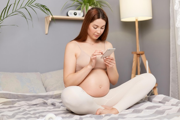 Piękna ciężarna brązowowłosa kaukaska kobieta siedzi w łóżku w pozie lotosu za pomocą telefonu komórkowego czytając nowe w sieciach społecznościowych wpisując wiadomości przeglądając internet