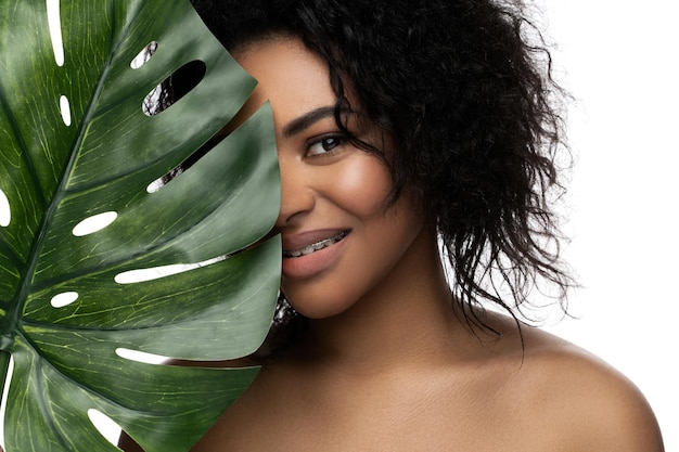 Piękna czarna kobieta o gładkiej skórze trzymająca zielony tropikalny liść na białym tle