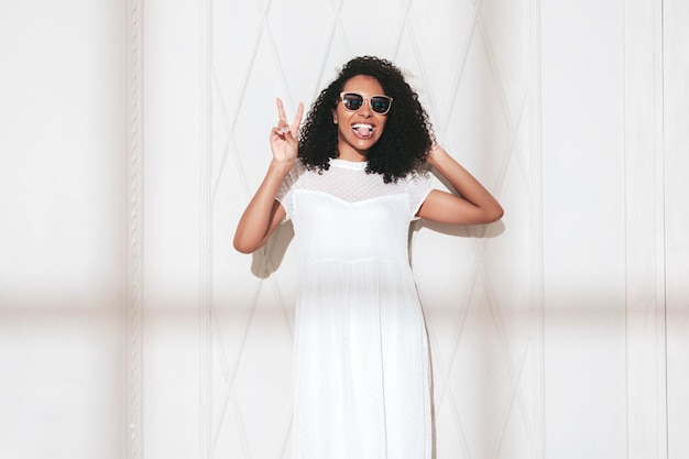 Piękna czarna kobieta z afro lokami fryzura Uśmiechnięta modelka ubrana w białą letnią sukienkę Seksowna beztroska kobieta pozuje przy ścianie w studio Opalona i wesoła W słoneczny dzień Cień z okna