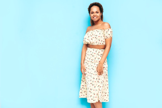 Piękna czarna kobieta z afro lokami fryzura Uśmiechnięta modelka ubrana w letnią sukienkę Seksowna beztroska kobieta pozuje przy niebieskiej ścianie w studio Opalona i wesoła
