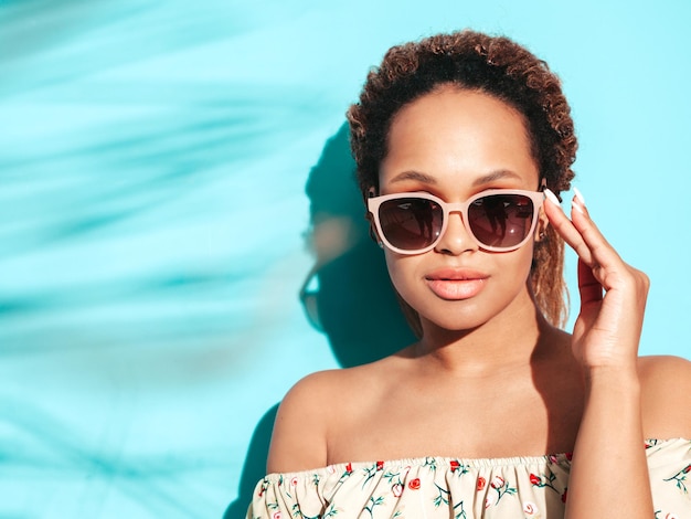 Piękna czarna kobieta z fryzurą afro loki Uśmiechnięta modelka ubrana w letnie ubrania hipster Seksowna beztroska kobieta pozuje przy niebieskiej ścianie w studio Opalona i wesoła w okularach przeciwsłonecznych