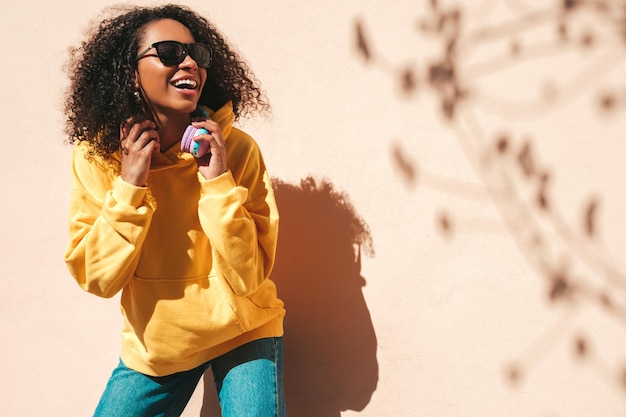 Piękna czarna kobieta z fryzurą afro lokiUśmiechnięta modelka hipster w żółtej bluzie z kapturem Seksowna beztroska kobieta pozuje na tle ulicy w pobliżu ściany w okularach przeciwsłonecznych Wesoły i szczęśliwy