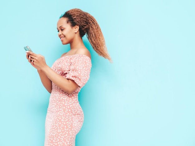 Piękna czarna kobieta z fryzurą afro lokiUśmiechnięta modelka w modnych letnich ubraniach Seksowna beztroska kobieta pozuje w pobliżu niebieskiej ściany w studioKorzystanie z aplikacji na smartfoniePatrząc na ekran telefonu komórkowego