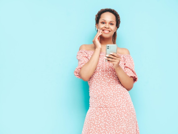 Piękna czarna kobieta z fryzurą afro lokiUśmiechnięta modelka w modnych letnich ubraniach Seksowna beztroska kobieta pozuje w pobliżu niebieskiej ściany w studioKorzystanie z aplikacji na smartfoniePatrząc na ekran telefonu komórkowego