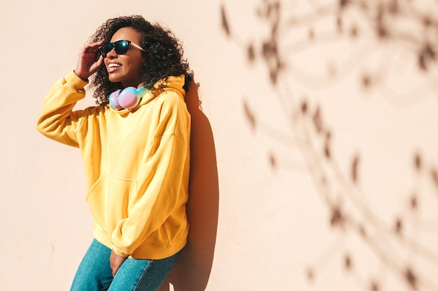 Piękna Czarna Kobieta Z Fryzurą Afro Lokiuśmiechnięta Modelka W żółtej Bluzie Z Kapturemseksowna Beztroska Kobieta Słuchająca Muzyki W Bezprzewodowych Słuchawkachpozowanie Na Tle Ulicy Przy ścianie W Okularach Przeciwsłonecznych