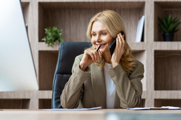 Zdjęcie piękna dojrzała bizneswoman siedzi przy biurku komputerowym w biurze i rozmawia przez telefon