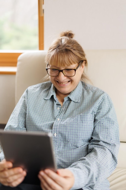 Piękna dojrzała kobieta w okularach używa cyfrowego tabletu i uśmiecha się siedząc na kanapie w domu