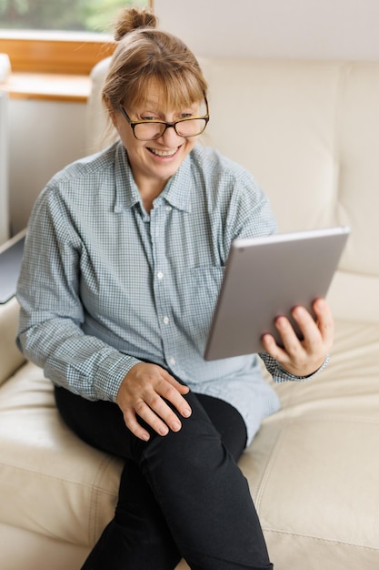 Piękna dojrzała kobieta w okularach używa cyfrowego tabletu i uśmiecha się siedząc na kanapie w domu