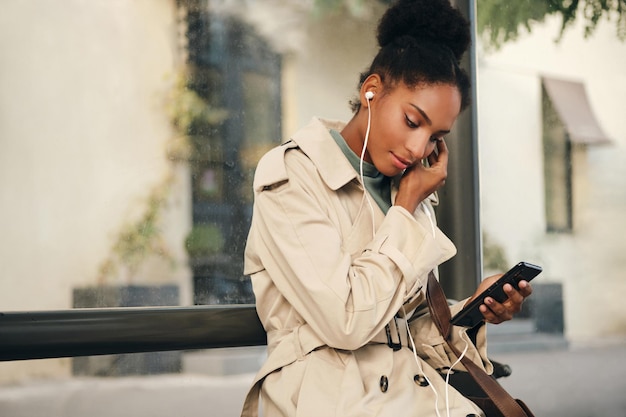 Piękna dorywczo Afroamerykańska dziewczyna w stylowym trenczu i słuchawkach, sennie słuchająca muzyki na telefonie komórkowym na przystanku autobusowym