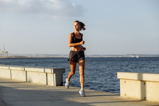 Piękna dziewczyna fitness w krótkich spodenkach i sportowej bluzce biegnie na nabrzeżu nad morzem podczas zachodu słońca