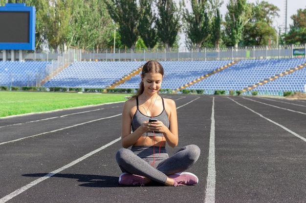 Piękna dziewczyna fitness w szarej odzieży sportowej używa smartfona i słucha muzyki na stadionie po treningu Sport i zdrowa koncepcja