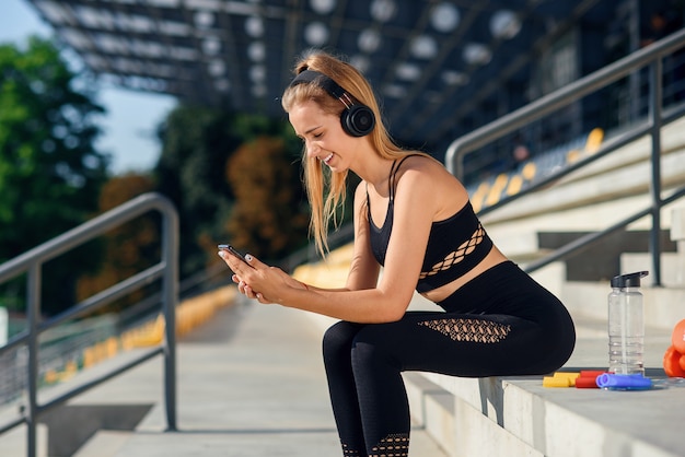 Piękna dziewczyna fitness w szarej odzieży sportowej używa smartfona i słucha muzyki na stadionie po treningu.