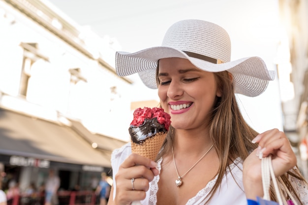 Zdjęcie piękna dziewczyna je lody na miasto ulicie z białym kapeluszem i torba na zakupy