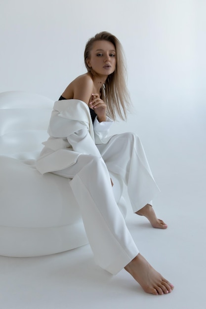 Piękna dziewczyna kaukaski w białym garniturze siedzi w pozie moda w studio na białym tle i patrzy w kamerę