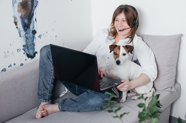 Piękna dziewczyna leży na kanapie w domu z laptopem w rękach, obok psa Jack Russell