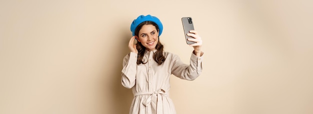 Piękna Dziewczyna Robi Selfie Na Smartfonie W Płaszczu I ładnym Kapeluszu, Uśmiechając Się Do Zdjęcia I Wyglądając Na Szczęśliwą
