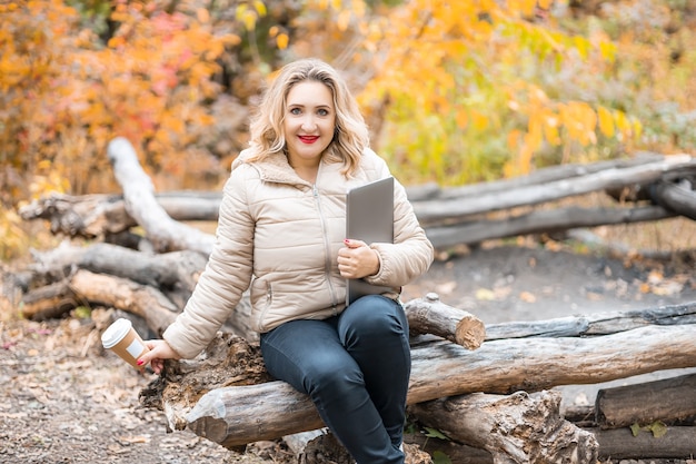 Piękna dziewczyna siedzi na kłodach w jesiennym parku, trzymając w dłoni laptopy i filiżanki kawy