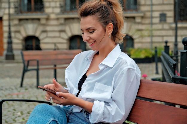 Zdjęcie piękna dziewczyna siedzi na ławce i trzyma telefon w dłoniach dziewczyna zamawia jedzenie przez telefon dziewczyna rozmawia przez telefon śmiejąc się szczęśliwa zdziwiona