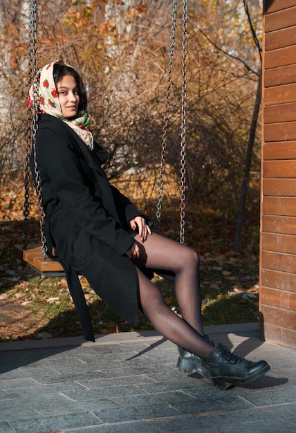 Piękna dziewczyna siedzi w parku na huśtawce na głowie to tradycyjna rosyjska chusta