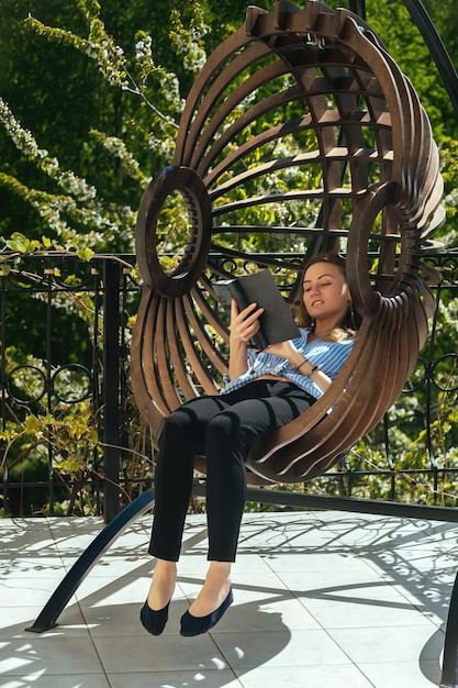 Piękna dziewczyna siedzi w wiszącym fotelu bujanym na tarasie w domu, relaksując się i czytając książkę