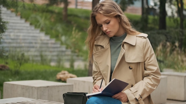 Piękna dziewczyna student szuka skoncentrowanego odrabiania lekcji na studia na ławce w parku miejskim. Stylowa blond dziewczyna studiująca na świeżym powietrzu