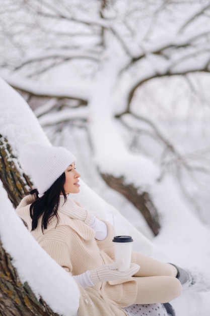 Piękna dziewczyna w beżowym swetrze i białym kapeluszu, ciesząca się piciem herbaty w śnieżnym zimowym lesie w pobliżu jeziora
