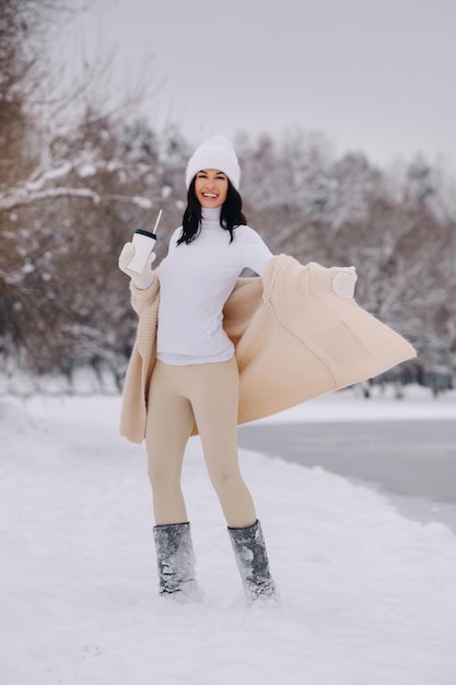 Piękna dziewczyna w beżowym swetrze i białym kapeluszu ze szklanką herbaty cieszy się śnieżnym nabrzeżem nad jeziorem
