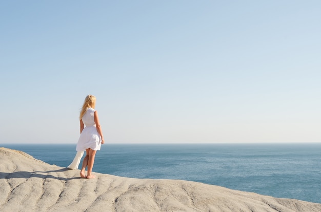 Piękna dziewczyna w białej sukni stojącej na skale i patrząc na morze
