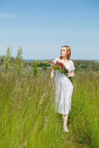 Piękna dziewczyna w białej sukni z kwiatami na polu z widokiem na niebo
