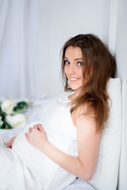 Piękna dziewczyna w ciąży siedzi na łóżku i uśmiecha się.