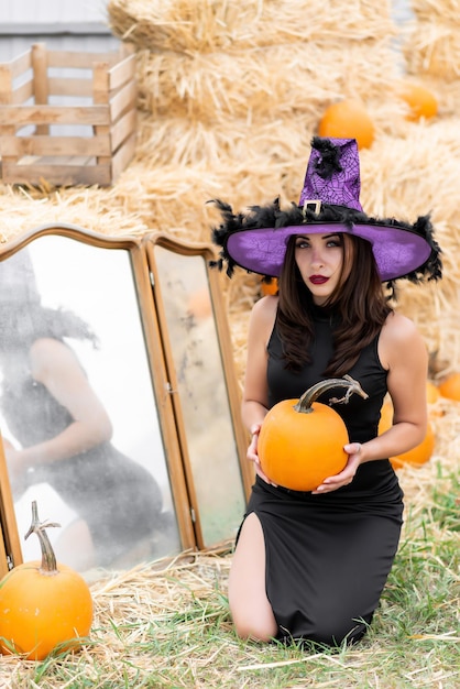 Piękna dziewczyna w czarnej sukience i kapeluszu wiedźmy pozuje przy starym lustrze Na tle siana Trzyma w rękach dynię Halloween dynia Wystrój z dyni
