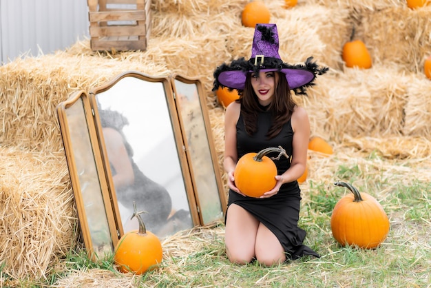 Piękna dziewczyna w czarnej sukience i kapeluszu wiedźmy pozuje przy starym lustrze Na tle siana Trzyma w rękach dynię Halloween dynia Wystrój z dyni