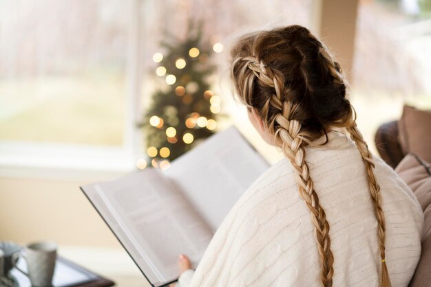 Zdjęcie piękna dziewczyna w jasnym pokoju bożonarodzeniowym czyta książkę i pije herbatę