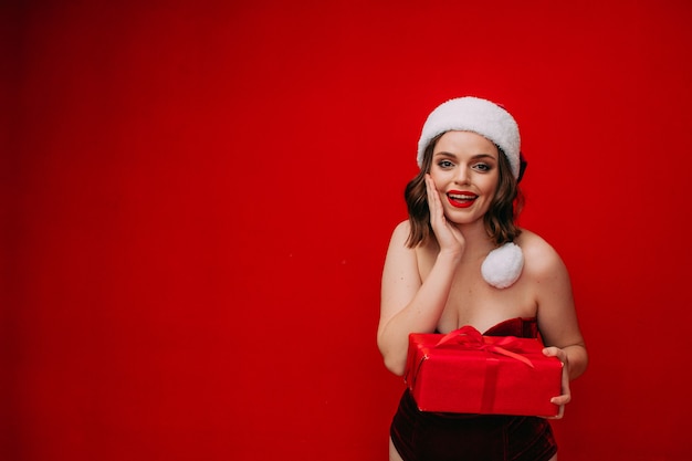 Piękna dziewczyna w kapeluszu Świętego Mikołaja trzyma w rękach prezent noworoczny na czerwonym tle