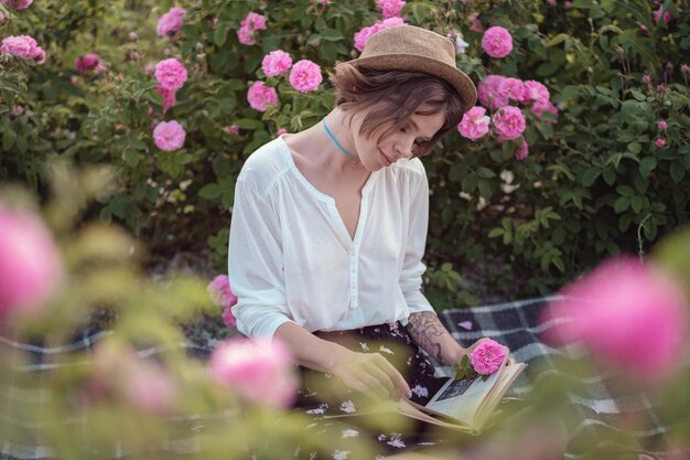 Piękna dziewczyna w kapeluszu z książką siedząca na trawie w różanym ogrodzie Wiejska dziewczyna cieszy się winem o zachodzie słońca i czyta książki w pachnącym ogrodzie różanym Koncepcja perfum