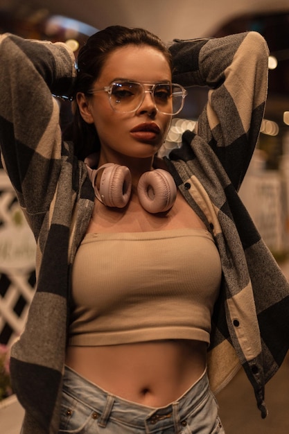 Piękna dziewczyna w modnych okularach i słuchawkach w stylowym topie z kraciastą koszulą stoi na nocnej ulicy
