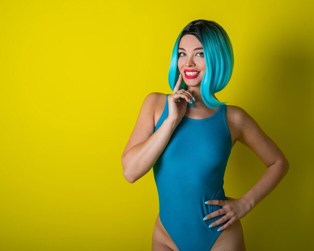 Piękna dziewczyna w niebieskiej peruce i jasnym bikini pozuje na żółtym tle Kobieta ze sztucznymi włosami i kostiumem kąpielowym podkreślającym pośladki