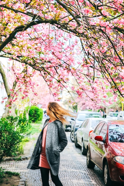 Piękna dziewczyna w pobliżu drzew sakura. Kobieta w sukience i stylowym płaszczu Różowe kwiaty kwitnące w Użhorodzie. Rozkwitaj wokół. Wiosna. Koncepcja relaksu i szczęścia.