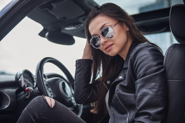 Zdjęcie piękna dziewczyna w skórzanej kurtce i okularach przeciwsłonecznych patrzy na kamerę, siedząc w samochodzie